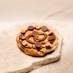 Cookie garni de pâte à tartiner, noisettes et pastilles de chocolat sur pierre claire.