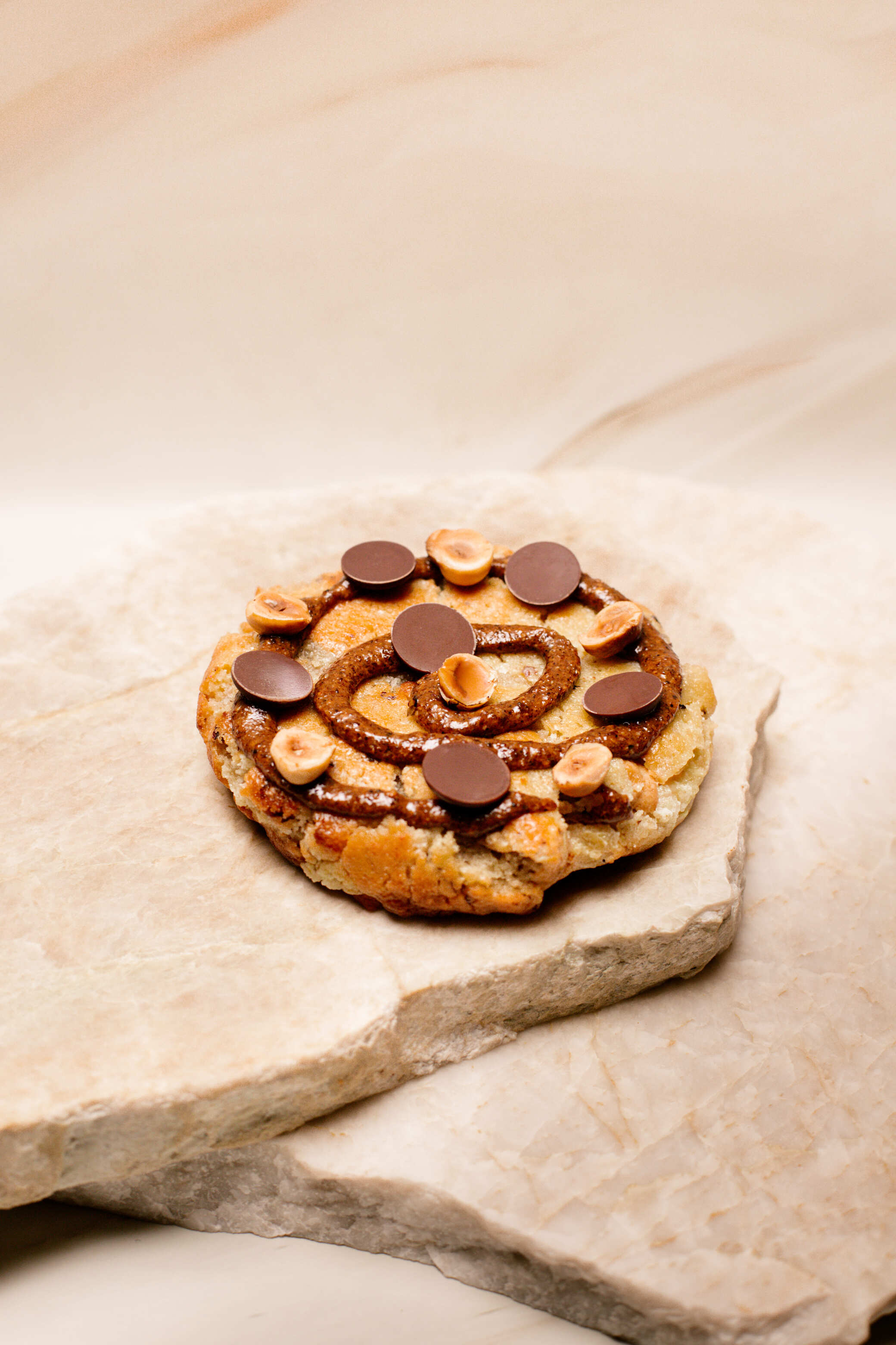 Cookie garni de pâte à tartiner, noisettes et pastilles de chocolat sur pierre claire.