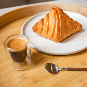Croissant doré sur assiette blanche avec café espresso en tasse en verre et cuillère sur plateau en bois.
