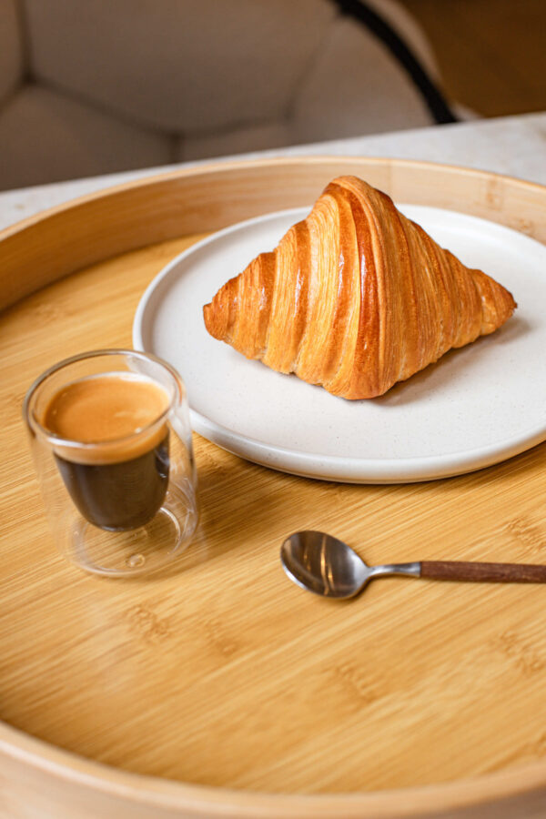 Croissant doré sur assiette blanche avec café espresso en tasse en verre et cuillère sur plateau en bois.