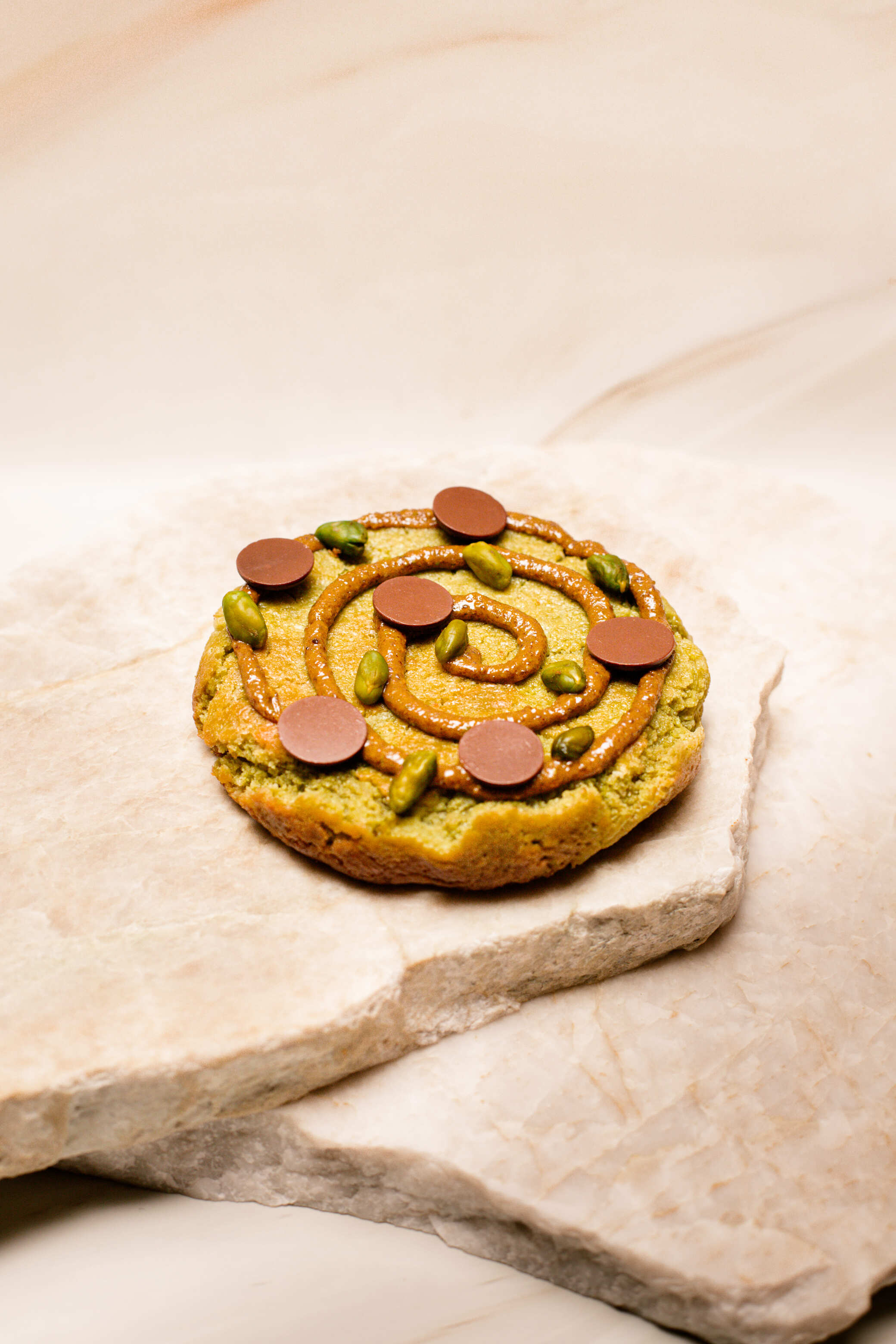 Cookie vert avec glaçage en spirale marron, pistaches et pastilles de chocolat sur pierres claires.