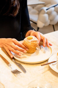 Personne tenant un gâteau rond sur une assiette blanche avec café et couverts sur table en marbre.