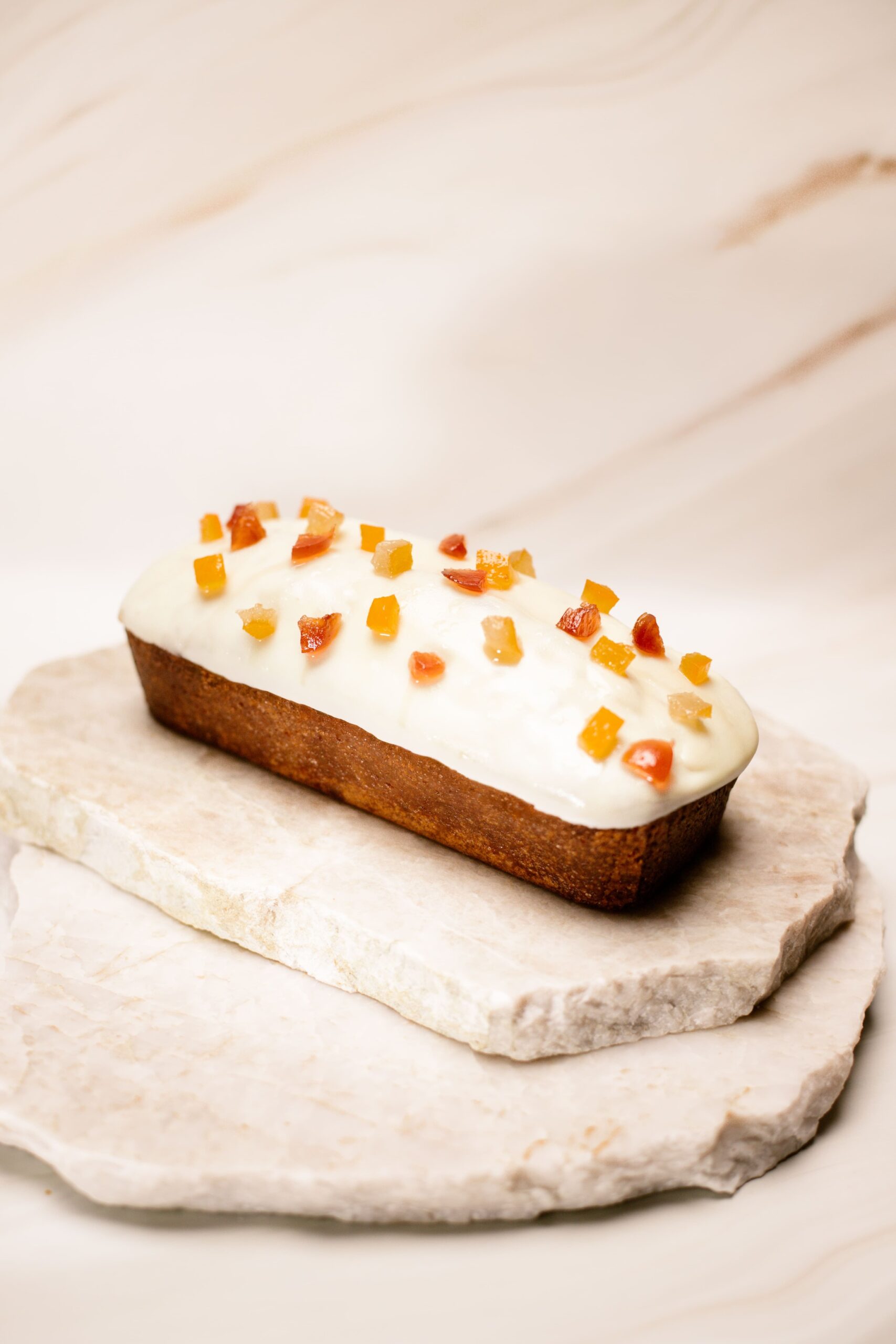 Cake rectangulaire glacé blanc avec morceaux de fruits confits sur une plaque en pierre.