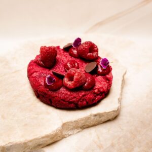 Dessert rond rouge garni de framboises, fleurs violettes, pastilles de chocolat sur pierre claire.