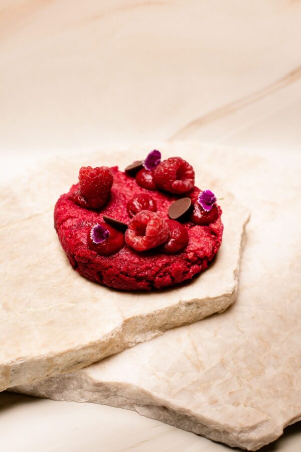 Dessert rond rouge garni de framboises, fleurs violettes, pastilles de chocolat sur pierre claire.
