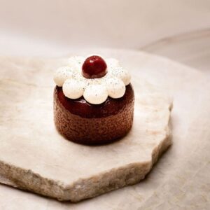 Petit gâteau rond au glaçage brillant avec fleur en crème blanche et cerise rouge sur pierre claire.