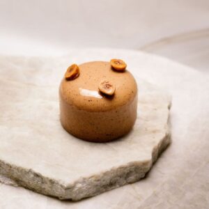 Pâtisserie ronde noisette glacée, décorée de trois noisettes entières sur plaques de pierre claire.