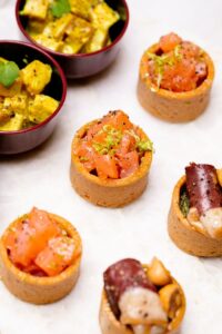 Verrines apéritives avec tartare de saumon, dés de fromage et charcuterie sur surface blanche.