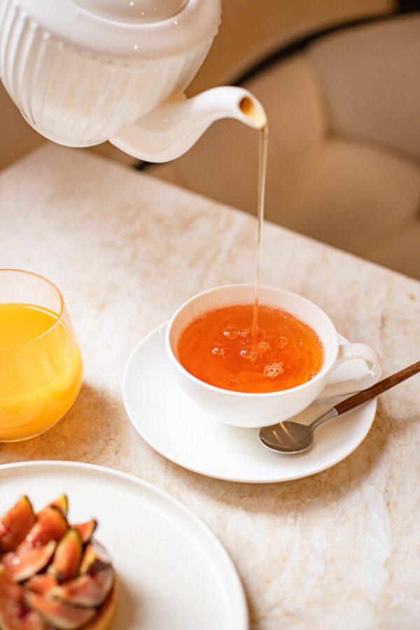 Versement de thé dans une tasse blanche avec un verre de jus d'orange et une pâtisserie aux figues.