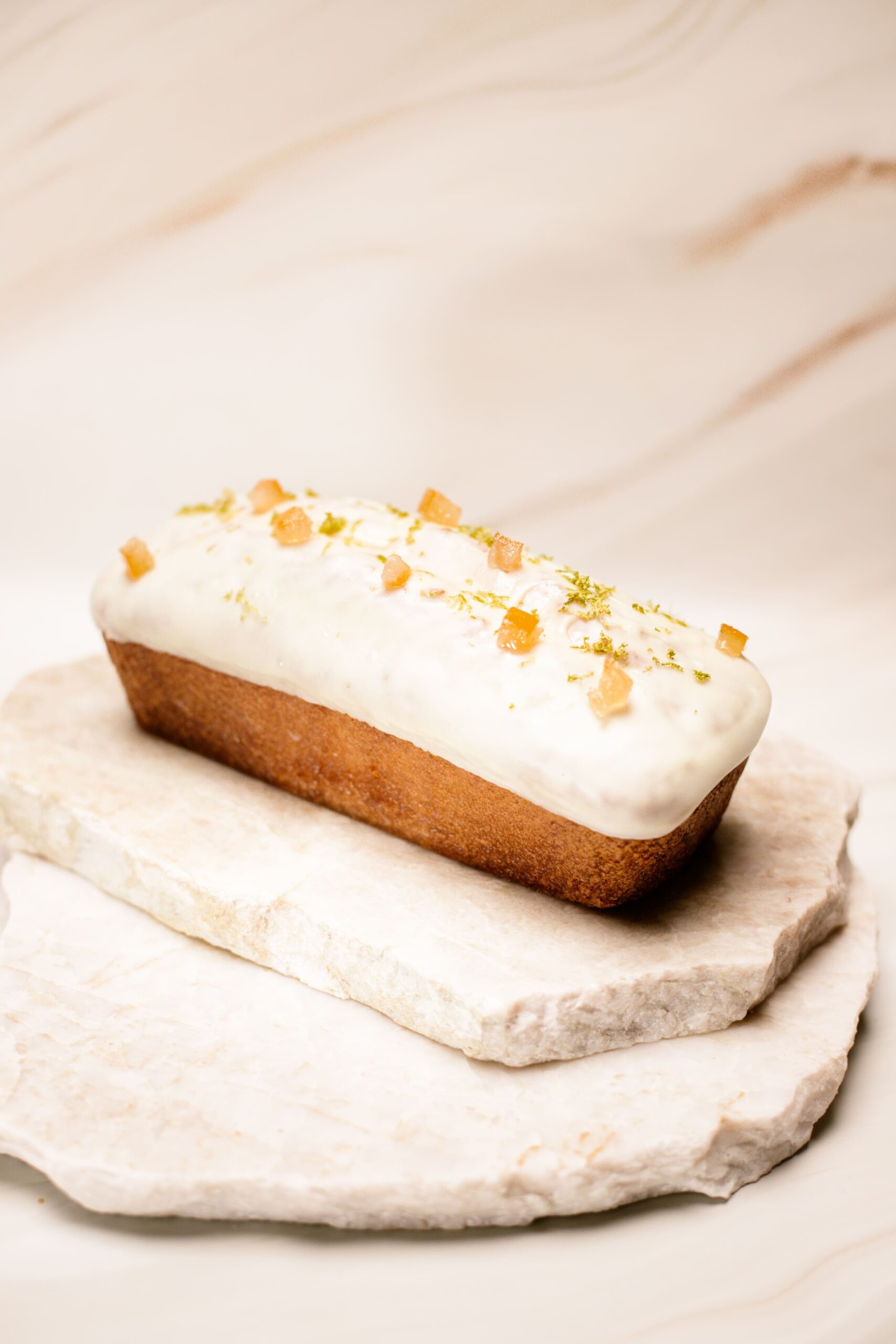 Gâteau rectangulaire glacé blanc décoré d'agrume confit et zestes verts sur pierres.
