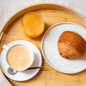 Plateau en bois avec café au lait, verre de jus d'orange et pain au chocolat sur assiette.