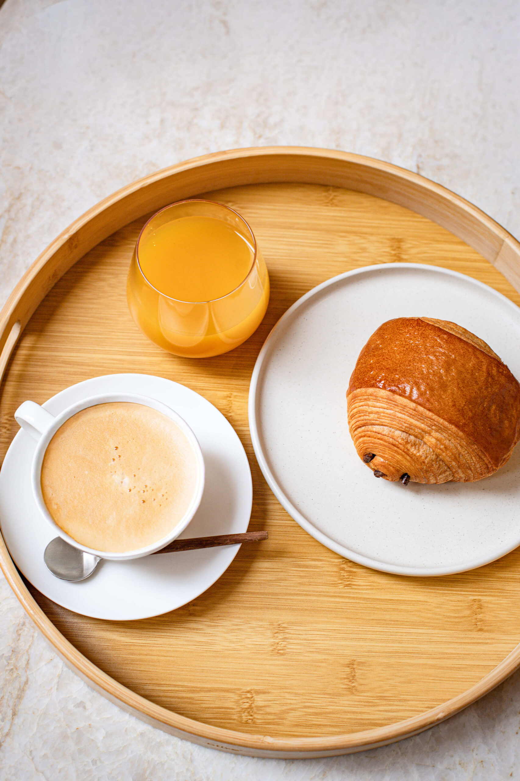 Plateau en bois avec café au lait, verre de jus d'orange et pain au chocolat sur assiette.