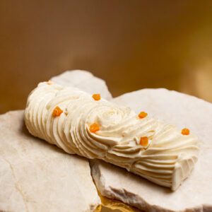 Dessert de Noël sans gluten en forme de bûche de couleur blanches avec des cubes oranges confits posé sur un marbre et un fond doré