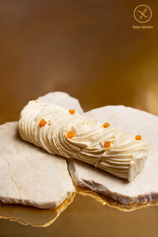 Dessert de Noël sans gluten en forme de bûche de couleur blanches avec des cubes oranges confits posé sur un marbre et un fond doré