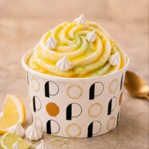 Pot de glace rond avec des lettres noires et dorées, rosace de glace pochée de couleur citron vert et jaune et de goût citron avec de meringues parsemées blanches.