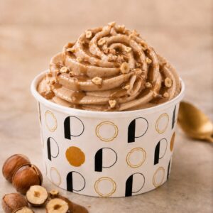 Pot de glace rond avec des lettres noires et dorées, rosace de glace pochée de couleur marron noisette et de goût noisette du Piémont avec des noisettes concassées.
