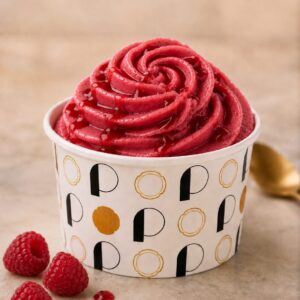 Pot de glace rond avec des lettres noires et dorées, rosace de sorbet pochée de couleur rouge framboise et de goût framboise avec un coulis framboise et des framboises fraîches découpées.